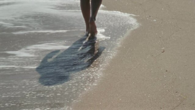 The feet of a girl going into the distance along the sandy shore of the sea washed by the waves