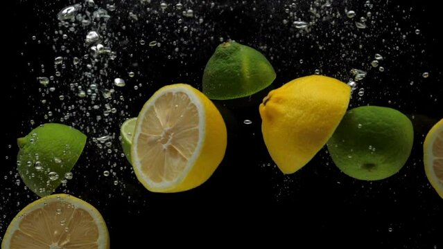 Slow motion close-up yellow lemon and green lime halves falling into transparent water on black background. Fresh fruits and berries splashing in aquarium. Citrus, healthy food, diet, air bubbles