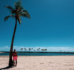 A man with a naked torso in orange shorts stands near a palm tree and looks at the ocean. Soft focus