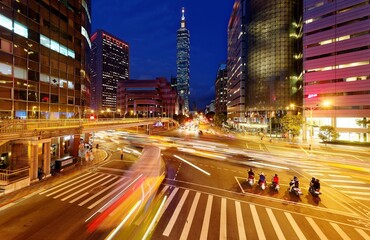 A bustling street corner in Downtown Taipei City with busy traffic trails at rush hour ~ Beautiful night scenery of Taipei 101 Tower & World Trade Center buildings in vibrant XinYi Financial District