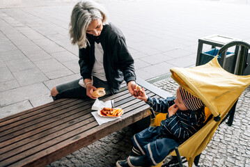 Woman sits on bench next to stroller with her son and eats curry wurst © Cavan
