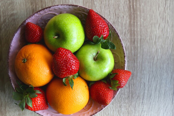 Pink bowl filled with fresh apples, oranges and strawberries on wooden table. Flat lay.