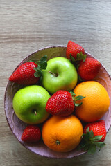 Pink bowl filled with fresh apples, oranges and strawberries on wooden table. Flat lay.