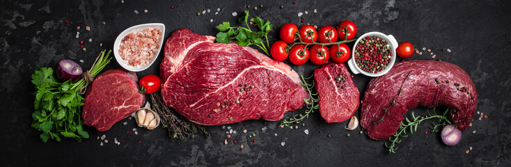 Assortment of raw cuts of raw beef meat steaks with spices on a dark background. Long banner...