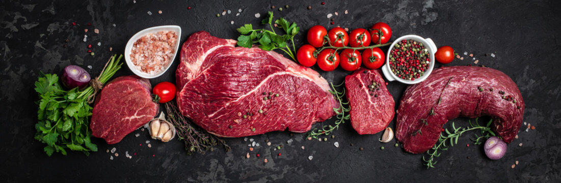 Assortment of raw cuts of raw beef meat steaks with spices on a dark background. Long banner format. top view