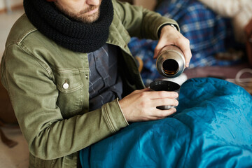 Close up of refugee pouring tea from thermos while hiding in shelter covered with sleeping bag and...