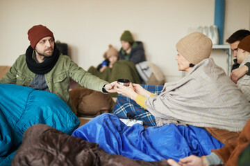 Group of Caucasian refugees sharing cup of tea while hiding in shelter together during war or...