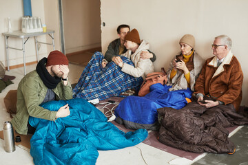 Group of distressed Caucasian refugees hiding in shelter during war or crisis covered with blankets...