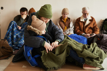 Side view of young mother holding sleeping son while hiding in refugee shelter and sitting on floor covered with blankets - obrazy, fototapety, plakaty