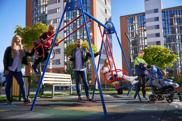 Young mum and dad swinging daughter and son, while other children standing near swing at playground. Side view of large family with four cute kids having fun together by swing set. Concept of holiday.