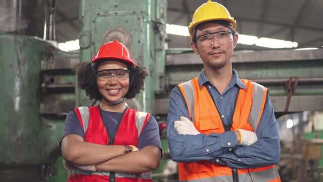 Zoom out shot of African American woman and Asian man factory worker in arms crossed looking smile at camera. Confident diversity mechanic or engineer standing in industrial construction site