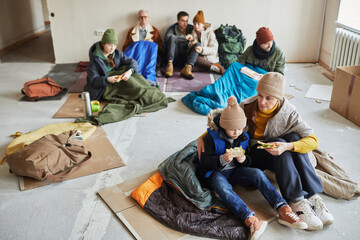 High angle view at group of Caucasian refugees eating food while hiding in shelter on floor covered...