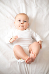 Portrait of a cute baby girl of 5 months in a white bodysuit on a white bed in the bedroom. Space for text. Child lifestyle