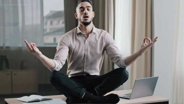 Mindful calm arabian business man employee in lotus position sit on work desk closed eyes meditating take break at home office keep mental balance patience yoga practices no stress feel zen balance