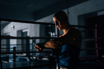 Fototapeta na wymiar Side view of serious bald boxer wrapping defense boxing tape around wrist before fight in sport club with dark interior on background of ring. Serious fighter getting ready for fight in fitness center