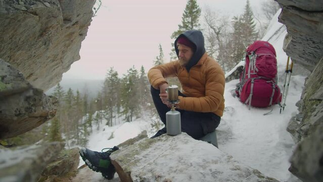 Man using mini portable gas stove while preparing hot tea in mountains while camping in winter