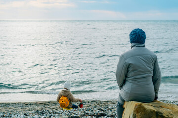 grandmother walk spring  sea, looking after her granddaughter playing. adult woman sits and looks at the autumn seascape, next to a child playing stones, healthy lifestyle, no gadgets, family weekend