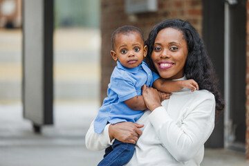 A beautiful African-American mom sitting on steps outdoors and holding her little boy and he looks...