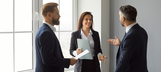 Team of three cheerful friends and coworkers talking during their break at work. Group of happy young people in formal suits enjoying a casual conversation while standing together by the office window
