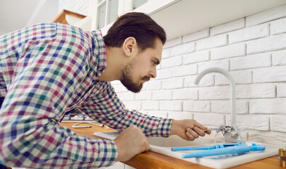 Plumber at work. Young repairman fixing the tap at home. Serious handsome man bending over the sink...