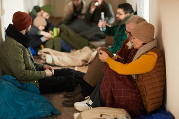 Side view portrait of senior refugees hiding in shelter during war or crisis and holding smartphone
