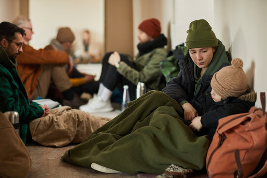 Caucasian young mother with child in refugee shelter sitting on floor and covered with blankets trying to keep warm