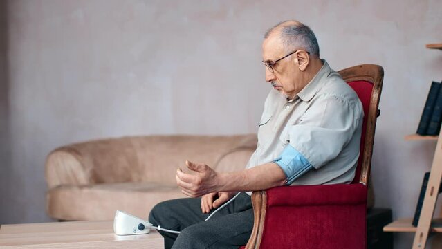 Senor man checking blood pressure use tonometer electronic device sitting red vintage armchair home