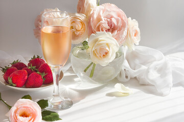 Bouquet of vintage romantic roses in pastel watercolor tones in a glass vase on a gray-white background of light and shadow. A glass of rose wine and strawberries, white silk 