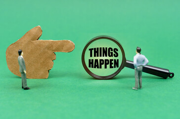 On a green surface, a hand, figurines of people and a magnifying glass with the inscription - Things Happen