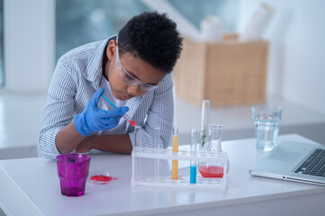 A dark-haired boy in a lab coat holding a syringe with a reagent and looking busy