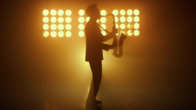 Silhouette jazz musician playing saxophone in spotlights. Man performing melody.