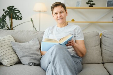 Middle-aged housewife sitting on sofa with book, free time retirement, hobby.