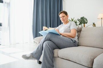 Attractive middle-aged woman with a lovely smile sitting on a sofa in the living room clutching a book.