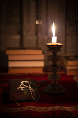 Still life with rosary on the prayer book, candle in vintage old candlestick and books near the window. Vintage old tablecloth on the table. Christianity concept. Time of prayer.