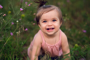 A baby in a pink romper with a ponytail smiles at the viewer from some long grasses with pink...