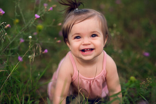 A baby in a pink romper with a ponytail smiles at the viewer from some long grasses with pink flowers.