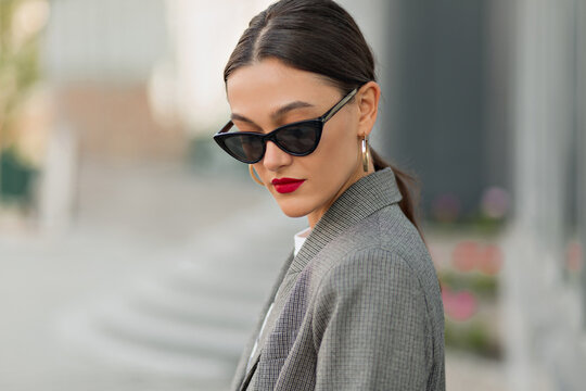 Portrait from back of elegant girl with dark hair is wearing gray jacket and sunglasses walking on street on glass building background
