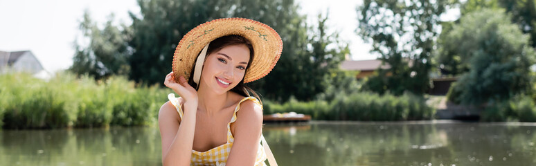 happy young woman in straw hat looking at camera near river, banner.
