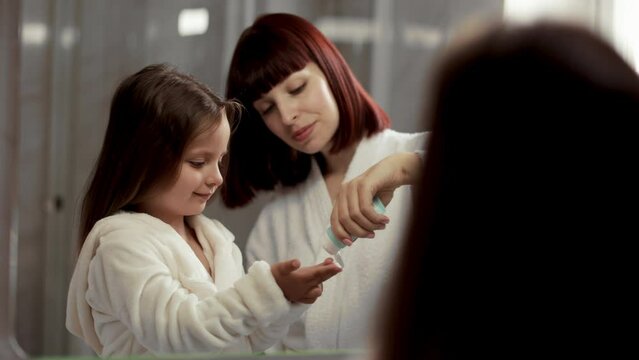 Mother squeezing treatment cream on theon hand of young and cute caucasian girl in white bathrobe standing in front of a mirror in the bathroom. The kid applying moisturizer on the cheeks.