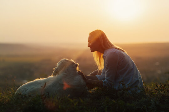 Happy pretty girl sitting on a lawn with a golden retriever dog at sunset. People with pets outdoors.