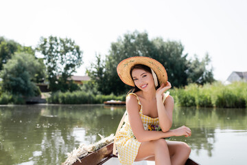 smiling young woman in straw hat and dress having boat ride on lake.