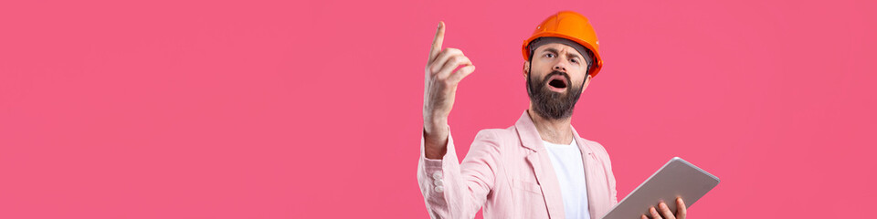 Portrait of young construction engineer wear orange hard hat, in a pink jacket standing on red studio background. A man with a tablet in his hands.