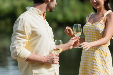 cropped view of happy young couple in summer clothes holding glasses with wine near lake.