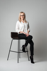 Beautiful young woman with glasses sitting on a chair in a studio with grey background. Sales agent, manager, agent, assistant, teacher, student