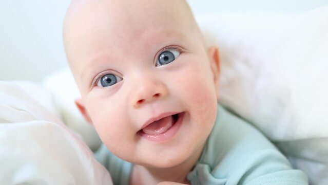 beautiful blue eyed infant baby portrait close-up, smiling face of little child lying in white bed on stomach. happy caucasian toddler baby portrait, funny kid looking camera opening mouth, emotions