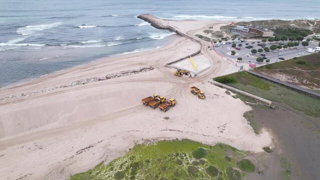 Esposende, Portugal, April 10, 2022: DRONE AERIAL FOOTAGE - The two sides of Restinga de Ofir. Esposende fishing harbor maintenance dredging. Dumper vehicles and rotating excavator parked on the sand.