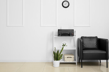 Shelf unit with modern printer, armchair and houseplant near light wall in room interior