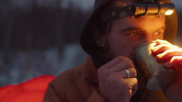 Chest up shot of bearded caucasian man with headlamp drinking hot tea from steel mug and looking in distance while standing outdoors at campsite in evening