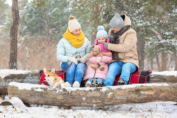 Little girl with toy, her grandparents and Corgi dog in forest on snowy winter day
