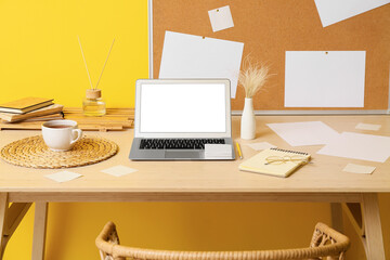 Modern workplace with laptop, sticky notes, vase and cup of tea near yellow wall